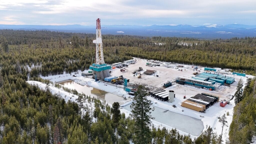 Aerial view of Mazama Energy’s drilling site at Newberry Volcano, Oregon—a tall rig, mud tanks and lined ponds on a snow-covered well pad surrounded by dense pine forest and distant mountains.